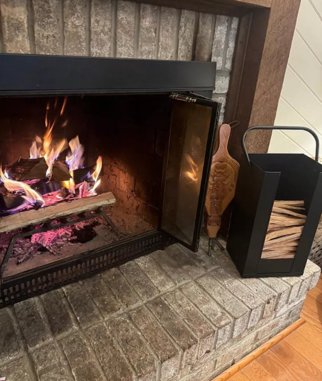 customer image of Cozy living room hearth with roaring fire, modern log holder filled with fire starter wood, and classic bellows tool