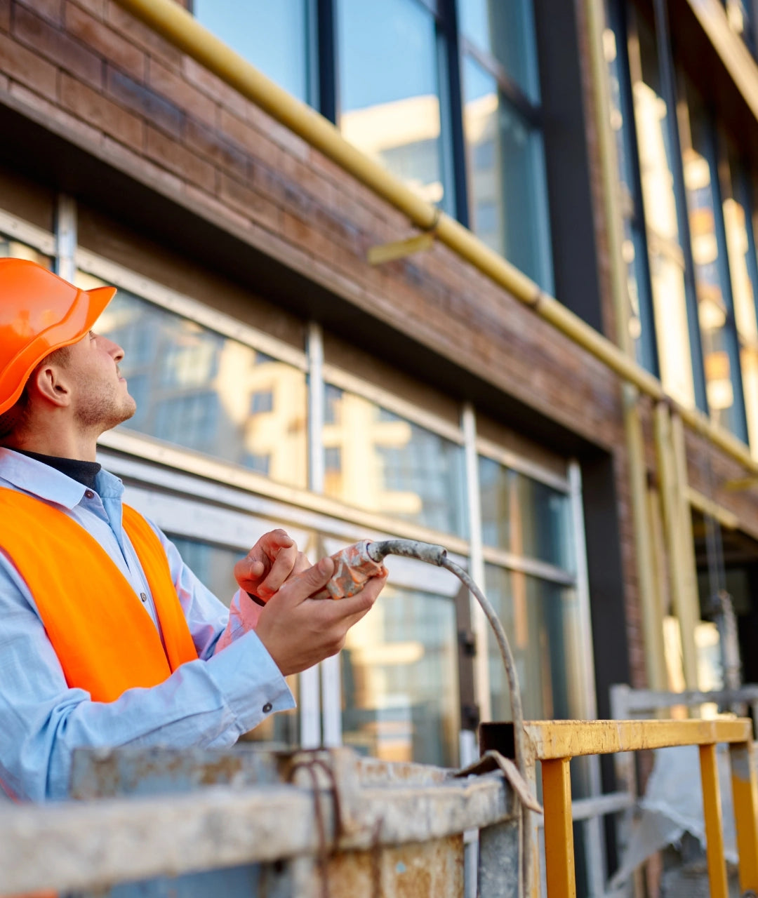 A construction worker in an orange hard hat and high-visibility safety vest stands on a scaffold near a modern building facade, holding a hose.