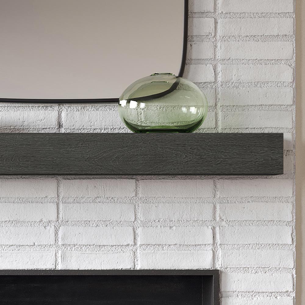 Alpine 60-inch Concrete Mantel Shelf in Ember finish, mounted on a white brick fireplace wall with a modern green glass vase and mirror above, showcasing its dark textured woodgrain design.