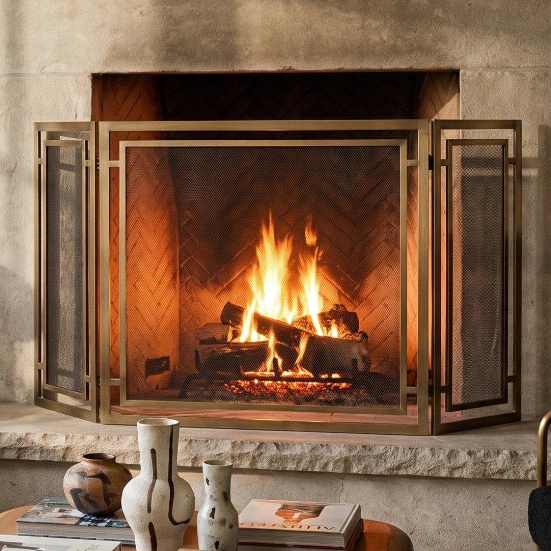 A brass Gatsby fireplace screen with glass panels stands open in front of a lit wood-burning fireplace, with a stone hearth and decorative vases in the foreground.
