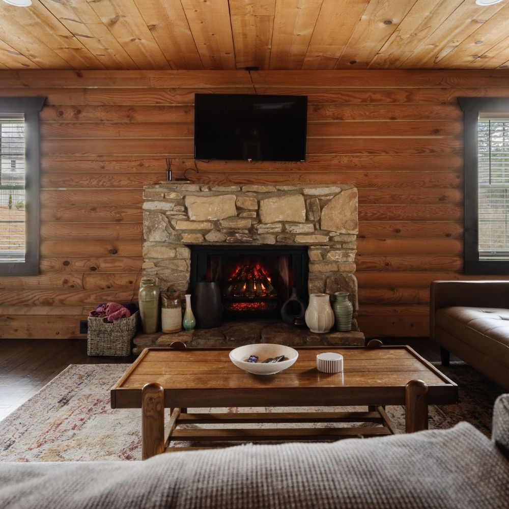 Modern Ember Free Stack Electric Log Set installed inside an existing non-working stone fireplace in a rustic log cabin Airbnb rental property. The warm living room features wood-paneled walls and ceiling, a natural stone fireplace surround, and a wood coffee table in the foreground. The electric log set glows with a realistic flame effect, surrounded by ceramic vases and decorative accents on the hearth. A wall-mounted TV sits above the mantel.