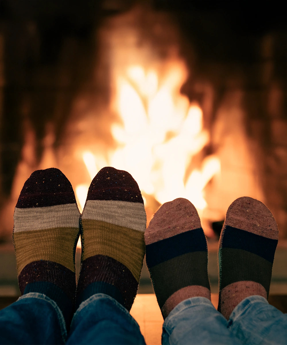 Two pairs of cozy striped socks worn on feet, with a warm fire burning in the background. The socks feature horizontal stripes in dark brown, beige, olive green, and navy blue, paired with blue jeans.