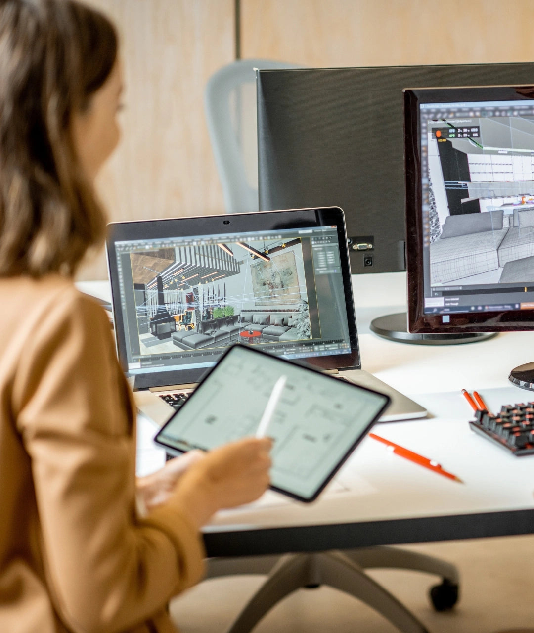 An interior design in a tan blazer sits at a desk using a tablet with a stylus, with a laptop and desktop monitor showing 3D interior design software in an office setting.