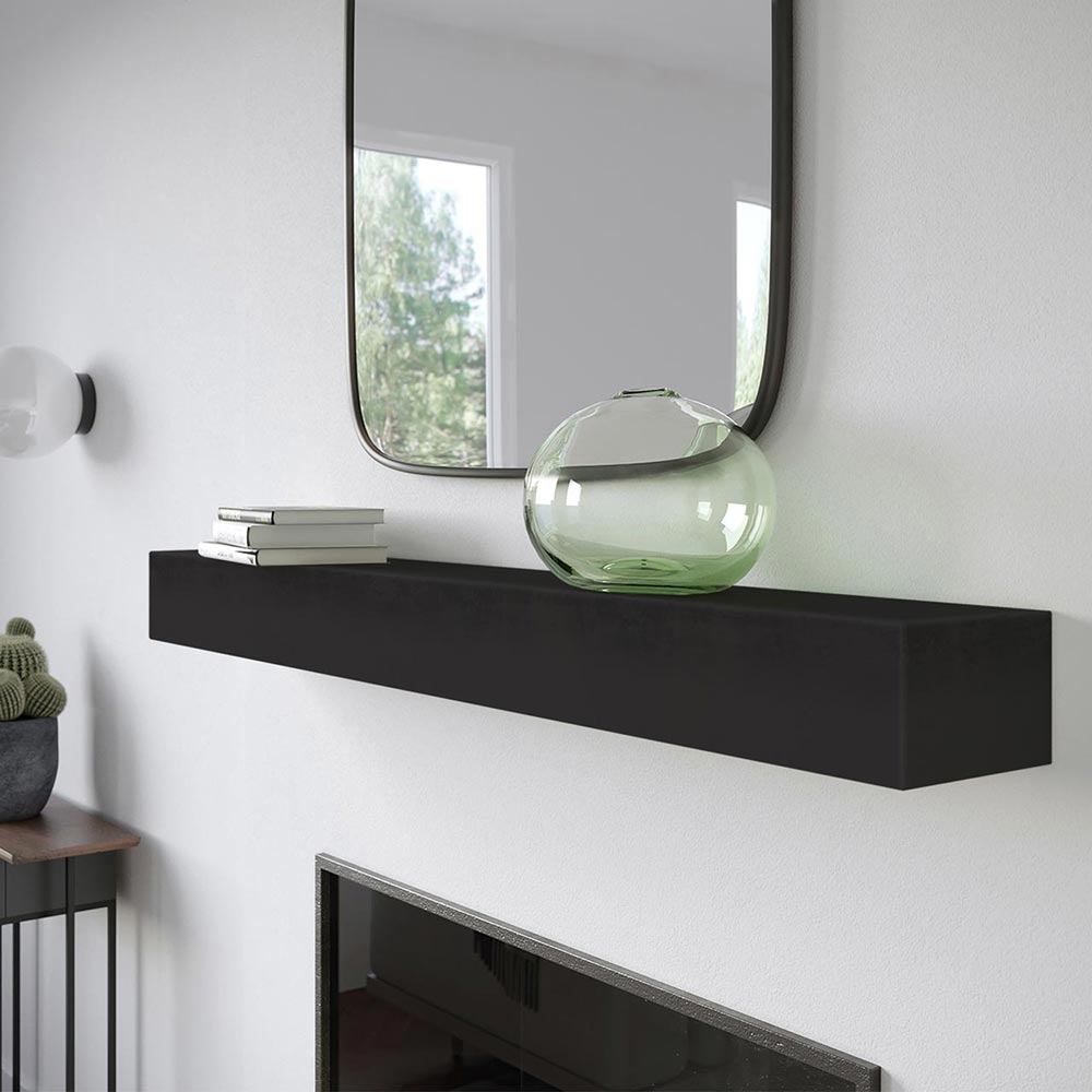 Angled view Everett 60-inch Concrete Mantel Shelf in Carbon finish, mounted on a white stucco fireplace with a square mirror, stacked books, and a green glass vase.