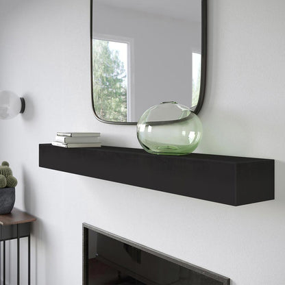 Angled view Everett 60-inch Concrete Mantel Shelf in Carbon finish, mounted on a white stucco fireplace with a square mirror, stacked books, and a green glass vase.