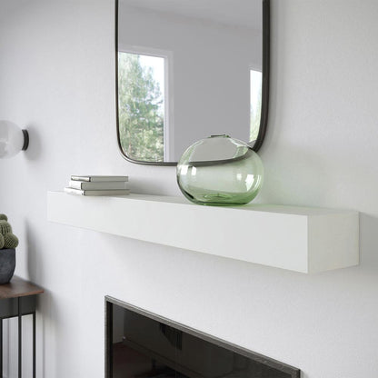 Angled view of Everett 60-inch Concrete Mantel Shelf in white Linen finish, mounted on a white wall fireplace with a square mirror, stacked books, and a green glass vase.
