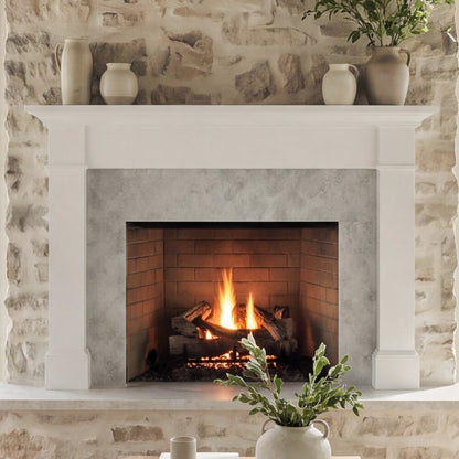 A white fireplace mantel and surround with a brick firebox and glowing fire inside, set against a rustic light stone wall with decorative vases and plants.