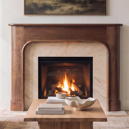 A warm fireplace scene with a dark wood mantel and frame, cream marble surround, glowing fire, and a light wood coffee table holding stacked books and a white bowl in a styled interior.
